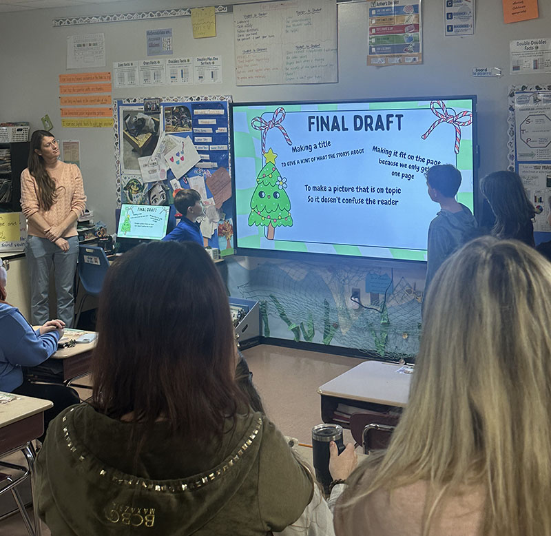 Fourth-graders present to a room of people. They are pointing to a slide they put together telling about the publishing a book.