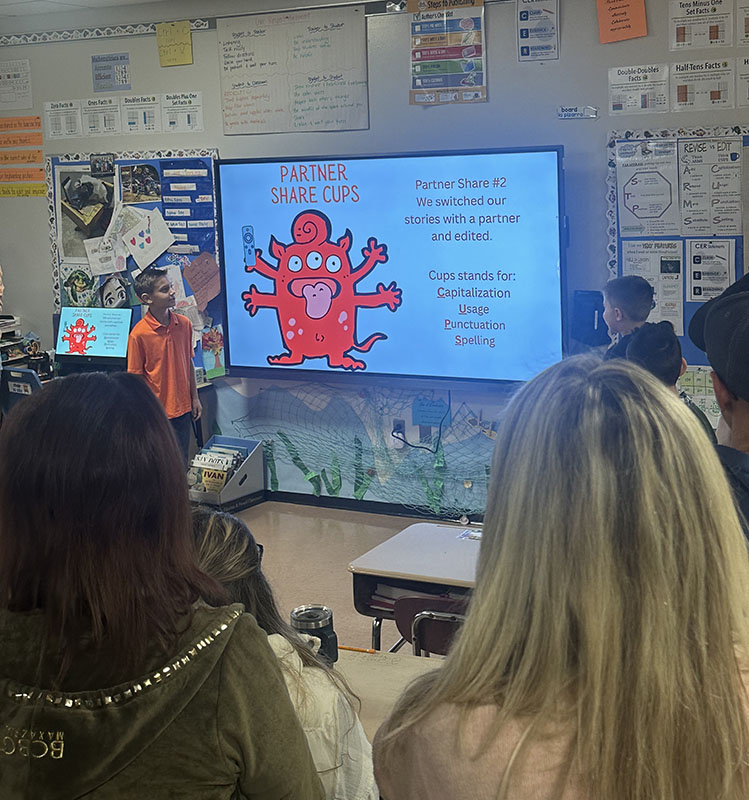 Fourth-graders present to a room of people. They are pointing to a slide they put together telling about the publishing a book.