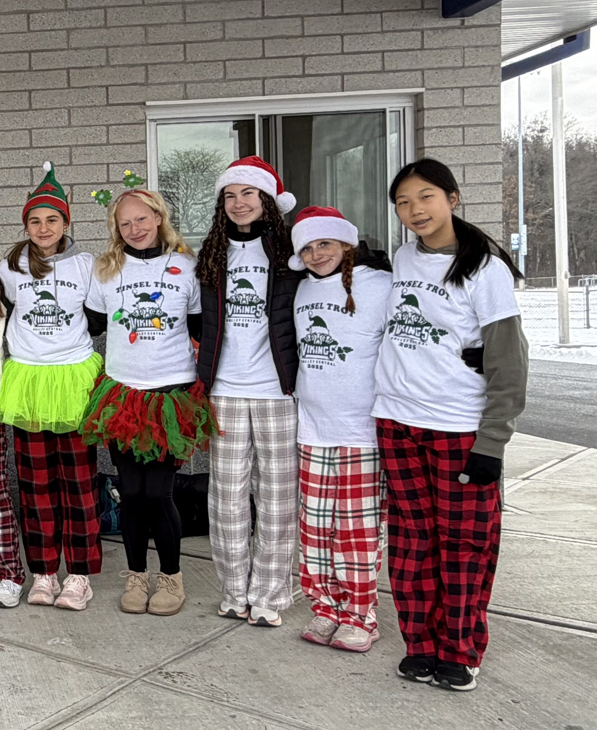 Five high school girls dress in various holiday attire- festive skirts and checked sleep pants, santa hats and elf hats. They are all wearing white tshirts that say tinsel trot.