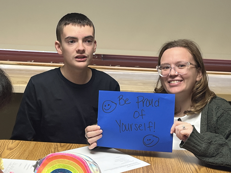 A woman with glasses smiles and holds up a blue piece of paper made by the student next to her.
