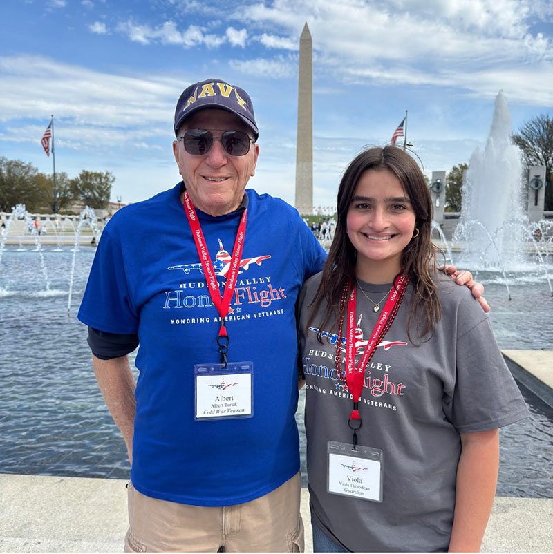 A younger woman and an older man stand by the reflecting pool with the Washington Monument behind them.