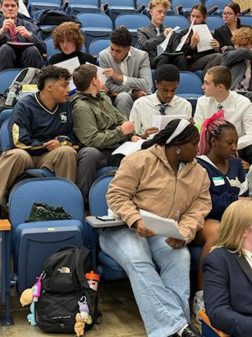 A large group of high school students sit in an auditorium talking toeach other.