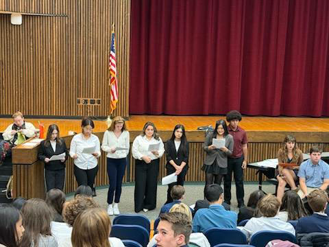 A group of 8 high school students stand in front of an auditorium filled with other students and present to them.