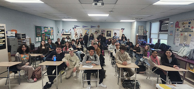 A group of about 45 students in a classroom. Many are sitting at desks and others are around the perimeter of the room. All are smiling.