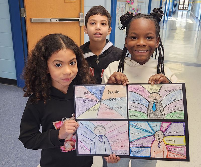 Three second-graders hold drawings of dr. martin Luther King Jr and the words to describe him.