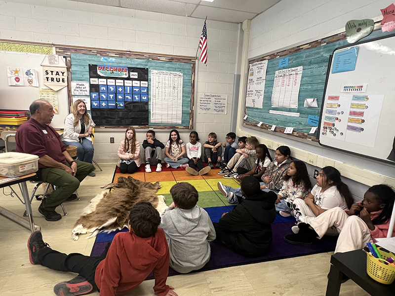 A class of fourth-graders sits around a colorful rug. A man sits at the top talking. 