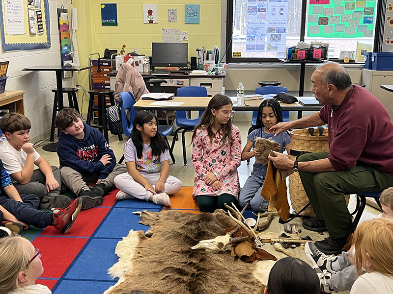 A man leans in to show elementary age kids a part of a deerskin.