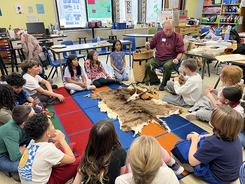 A man in a red shirt talks to a large group of fourth-graders who are sitting around a colorful rug.