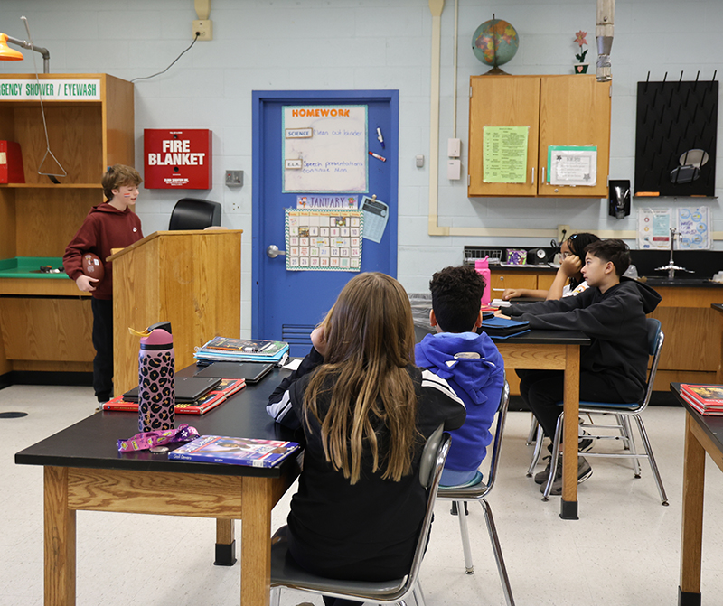 A middle school student stands at a podium speaking to her class.