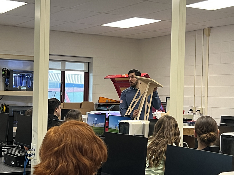 A man holds up a bench the class made. He is showing a group of middle school kids.