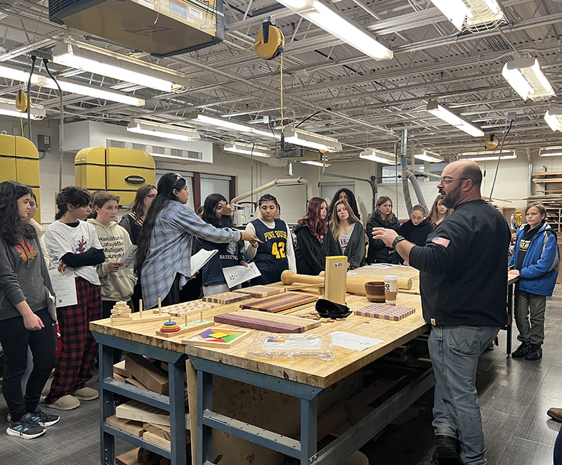 A teacher stands behind a woodworking table talking to middle school kids.