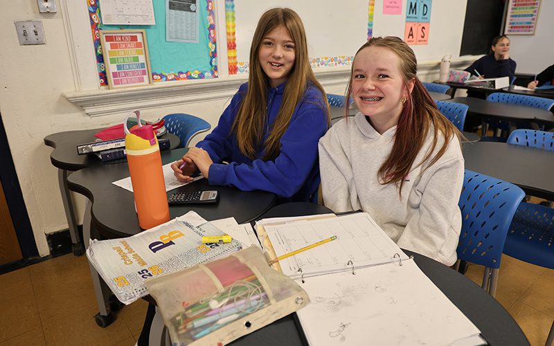 Crispell math smiling girls Two middle school girls sit at desks with books open and a calculator. They are smiling.