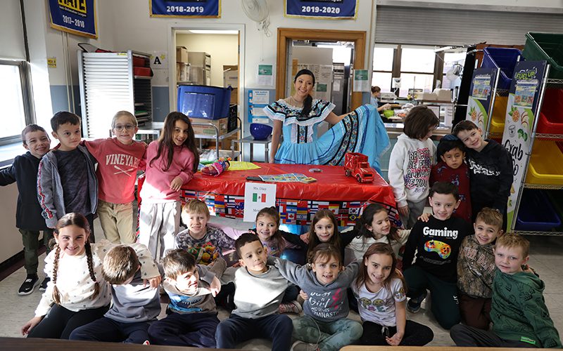 A woman stands with a class of young elementary students. She has a big blue Mexican dress and is spreading it out.