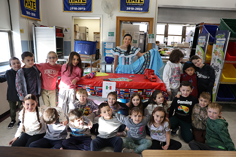 A woman stands with a class of young elementary students. She has a big blue Mexican dress and is spreading it out.