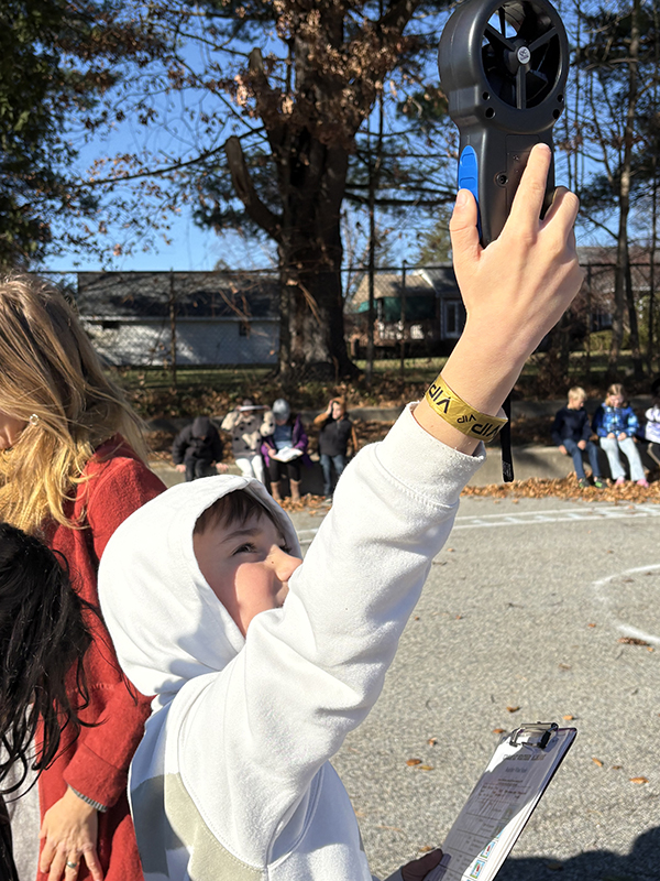 A third grade student, wearing a white hoodie, holds up a small device that measures wind speed.