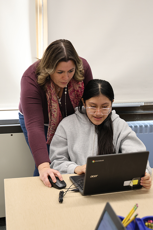 A woman with long dark hair leans over to help a fifth-grade student who is working on a Chromebook.