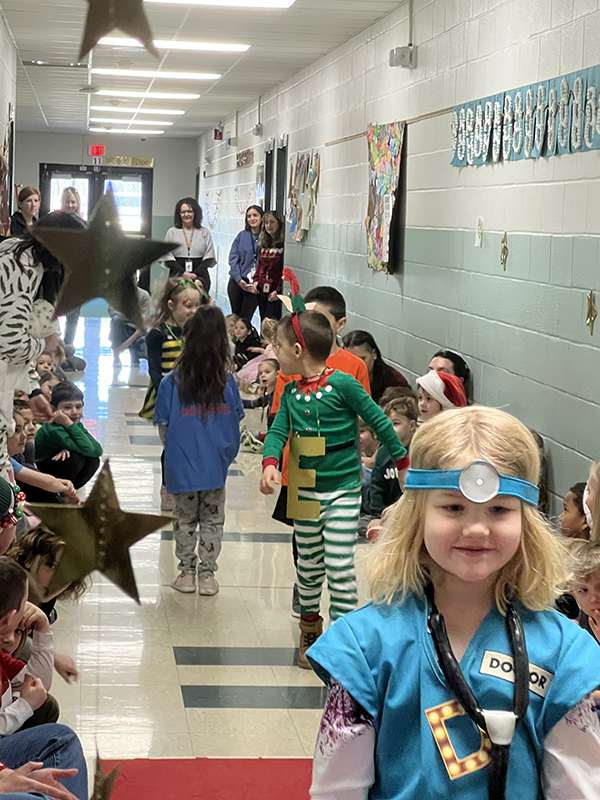 A group of kindergarten students march down a hallway with many students sitting on the sides.