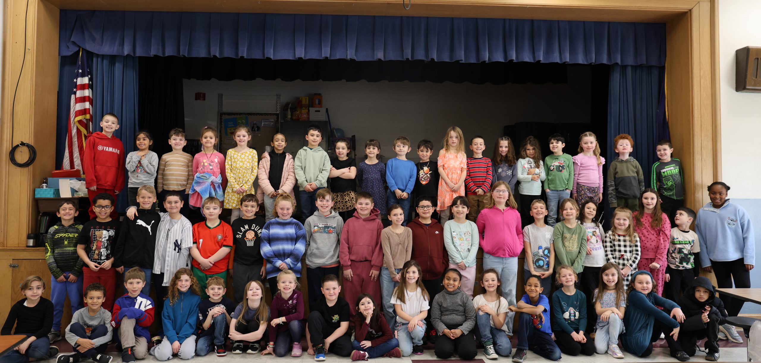 Three large rows of elementary kids stand on or near a stage. They are smiling.