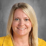 A woman with long blonde hair smiles. She is wearing a yellow shirt.