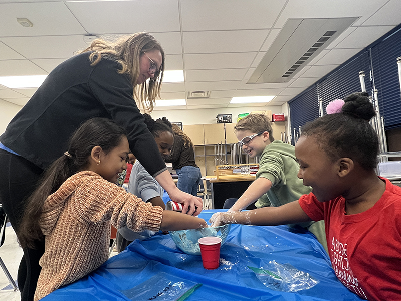A teacher stands over a table with four second-graders working at it. She pours baking soda and sprays shaving cream together to make snow. The kids have their hands in the mixture.