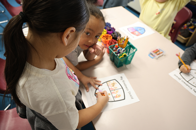 Two kindergarten students work at a table. One is coloring and the other is looking at the camera.