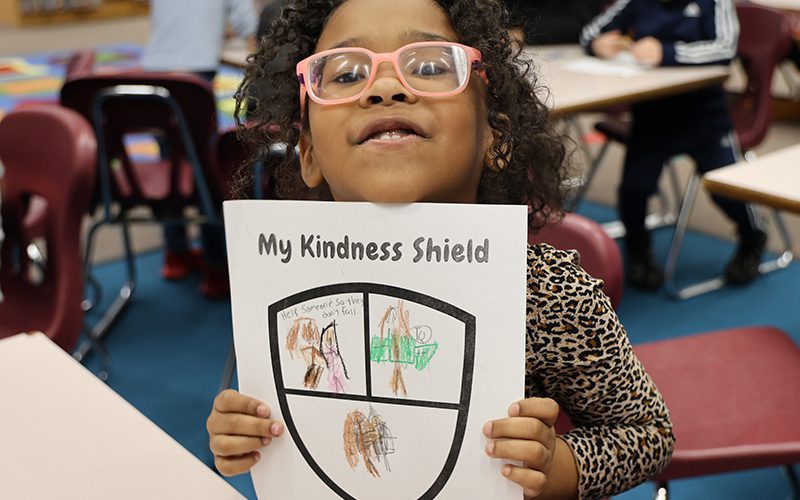 A kindergarten girl smiles and holds up a piece of paper where she colored a shield.