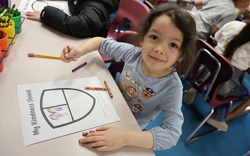 A kindergarten girl smiles. She is working on coloring a shield.