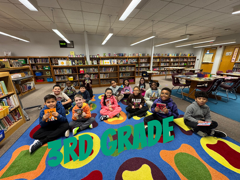 A group of elementary kids sit on a colorful rug and hold books.