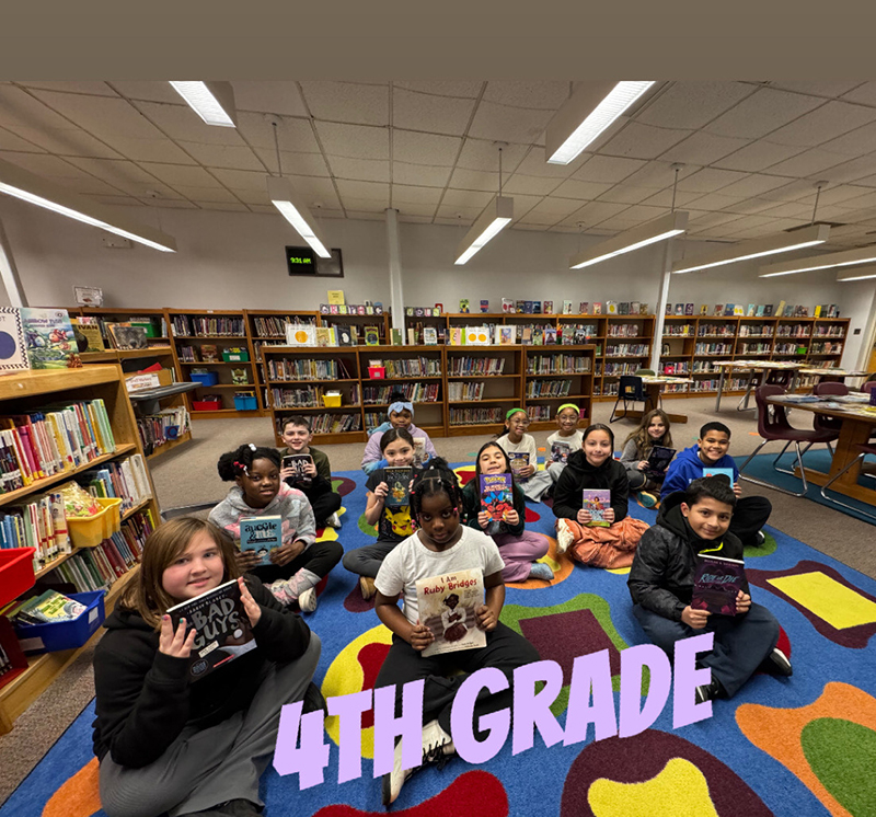 A group of elementary kids sit on a colorful rug and hold books.