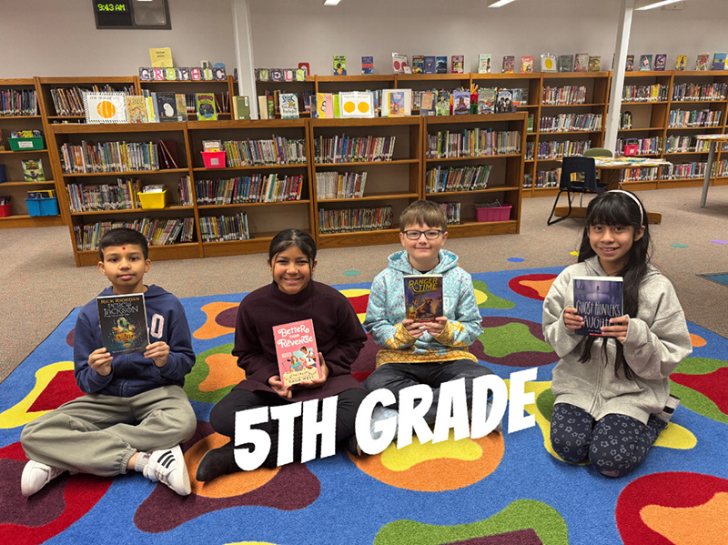 A group of elementary kids sit on a colorful rug and hold books.