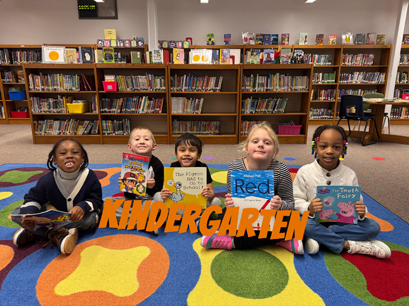 A group of elementary kids sit on a colorful rug and hold books.