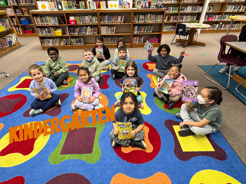 A group of elementary kids sit on a colorful rug and hold books.