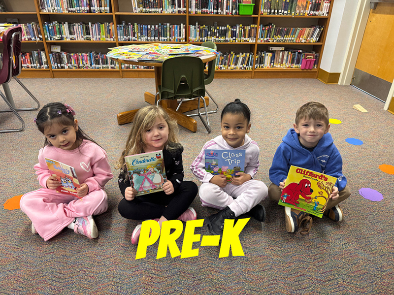 A group of elementary kids sit on a colorful rug and hold books.