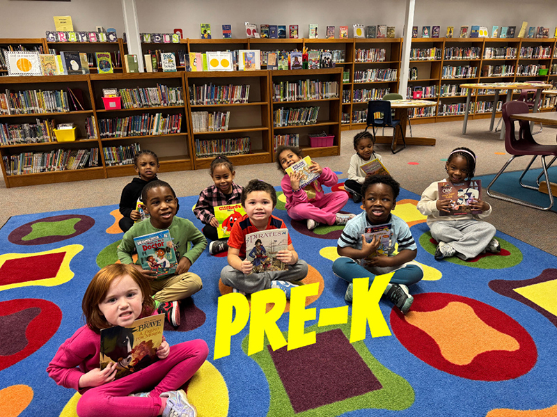 A group of elementary kids sit on a colorful rug and hold books.