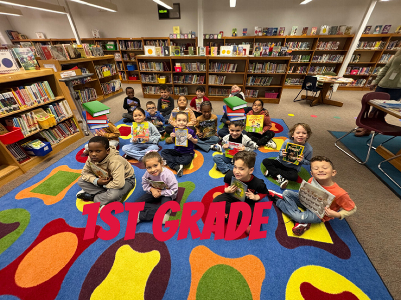 A group of elementary kids sit on a colorful rug and hold books.