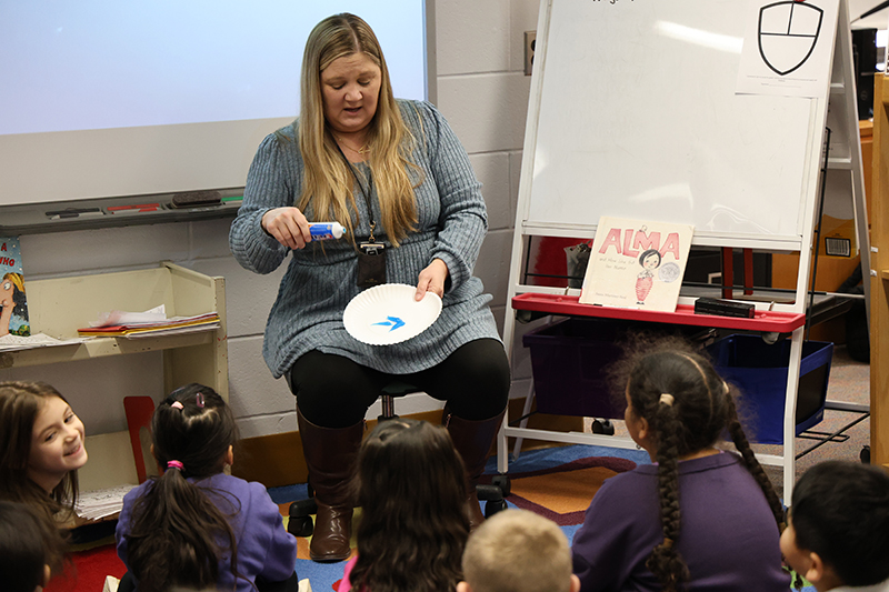 A teacher with long blonde hair squeezes blue toothpaste onto a plate while kindergarten students watch.