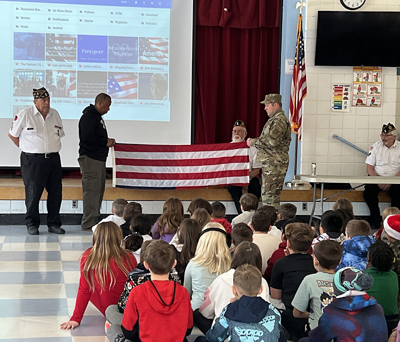 A group of elementary students sit on a floor and watch as two men fold an American flag. 