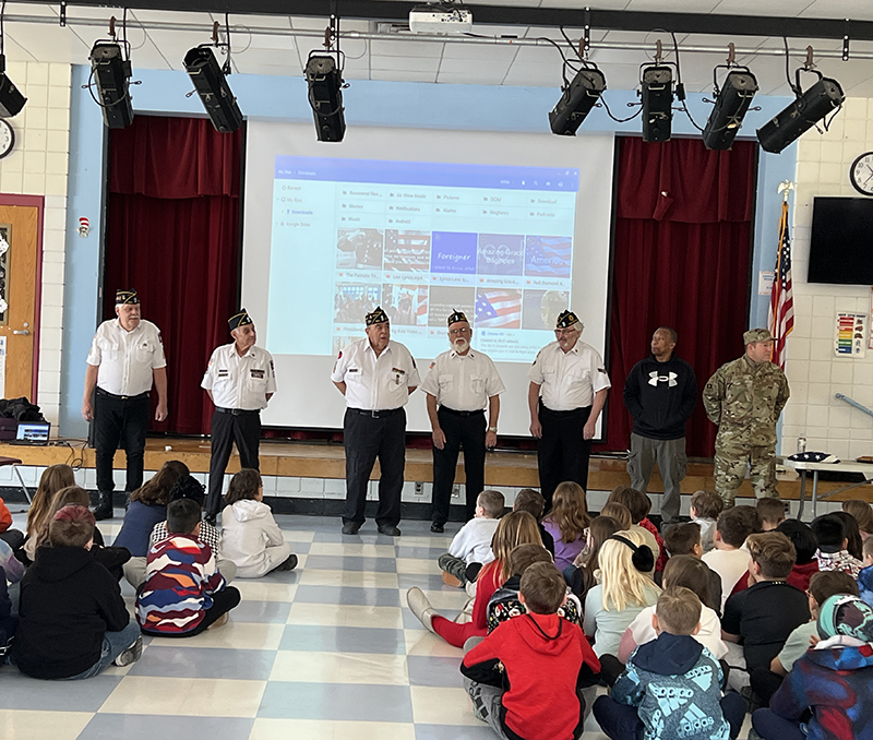 A group of even older men stand in front of a group of elementary students who are sitting on the floor. The men are wearing dark pants, white shirts and American Legion caps.