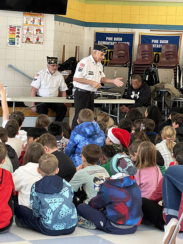 An older man dressed in a white shirt, blue pants and American legion cap talks to kids.