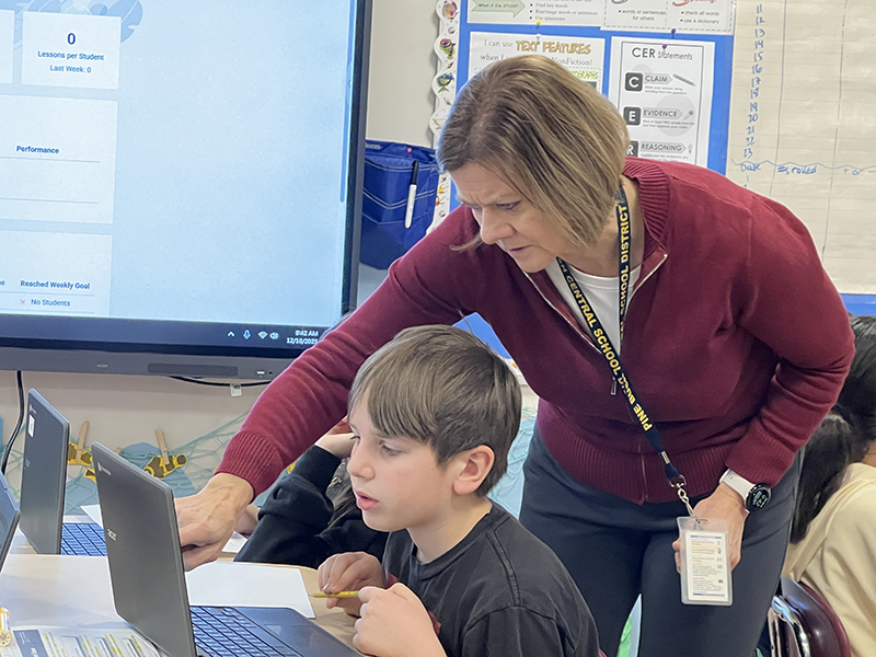 A woman with chin-length hair, wearing a cranberry shirt, points to something on a student's screen as he watches.