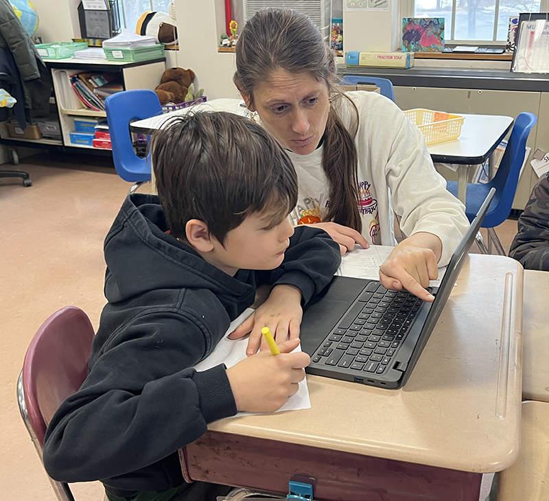 A woman with long hair in a ponytail over her shoulder points to  something on a Chromebook screen while helping a little boy.