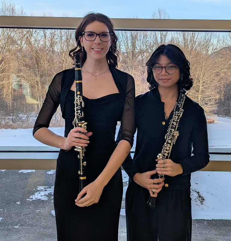 Two high school girls wearing black dresses and holding flutes stand together smiling.