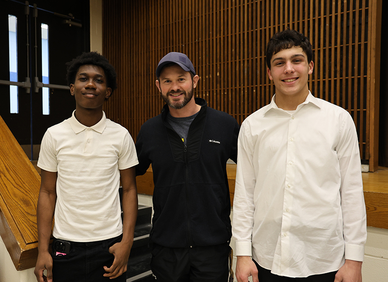 A man stands in the center and a young man stands on either side of him. They are smiling.