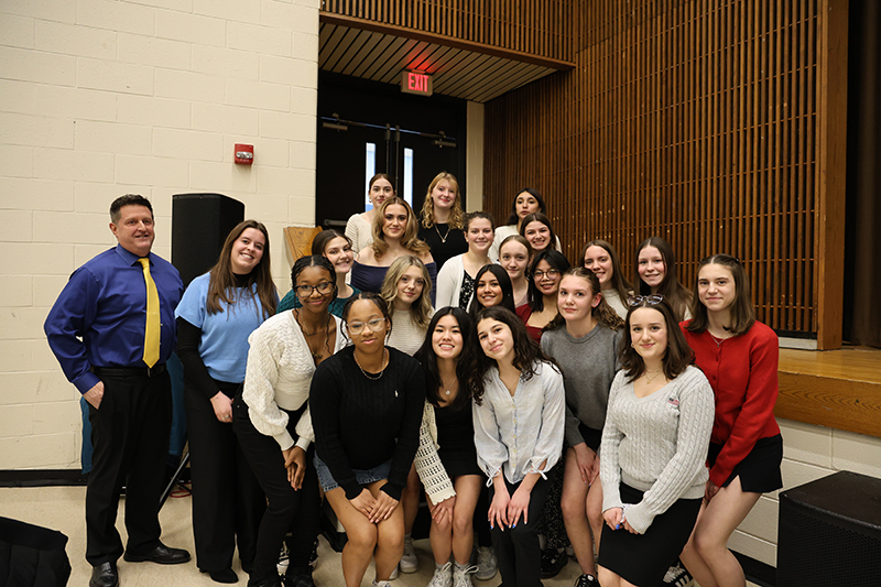 A man stands on the left with a large group of young women, who are standing together with some going up a short flight of stairs.