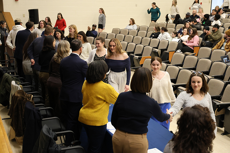A line of young women shake hands with a line of men and women behind a table.