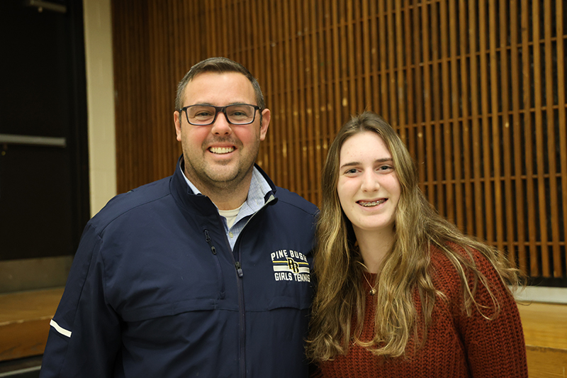 A man wearing glasses and a navy blue zip shirt smiles as he stands with a young woman with long hair, also smiling.