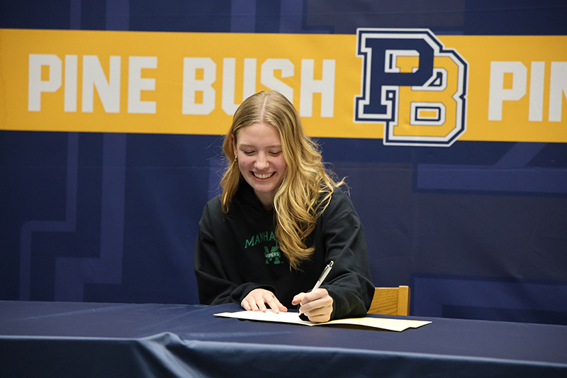 A high school girl with long blonde hair sits at a table, smiles and signs a piece of paper. Behind her is a banner that says Pine Bush PB.