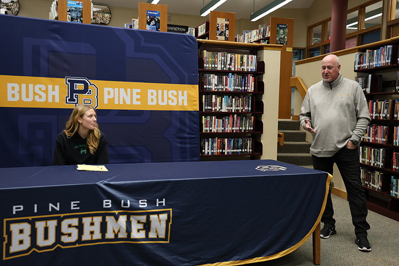 A man stands to the right and speaks, as a young woman sits at a table and smiles.
