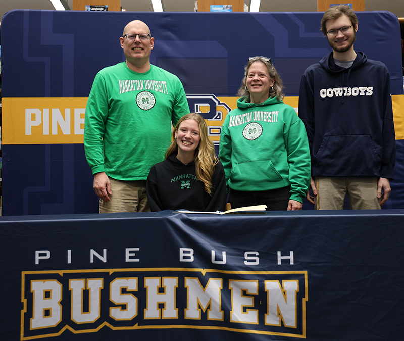 A high school senior with long blonde hair sits smiling at a table. Behind her are her family, two men and a woman. Everyone is smiling