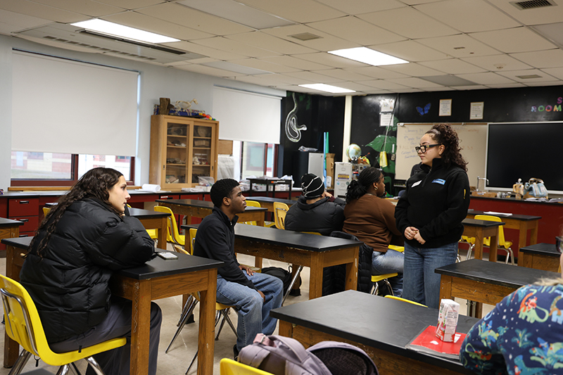 A woman stands amongst high school students who are sitting at desks as they talk.